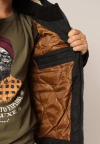 Veste extérieure noire avec un intérieur matelassé marron. Ferme par zip et possède un col côtelé. Poche intérieure visible, montrant un design épuré.