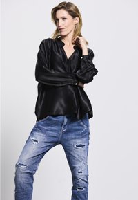 Schwarze Satinbluse mit langen Ärmeln und V-Ausschnitt, kombiniert mit ausgebeulten blauen Jeans aus Denim, die Risse und Farbspritzer aufweisen.
