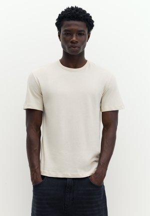 Jeune homme à la peau foncée et aux cheveux bouclés portant un t-shirt beige uni à manches courtes et un pantalon foncé, les mains dans les poches.