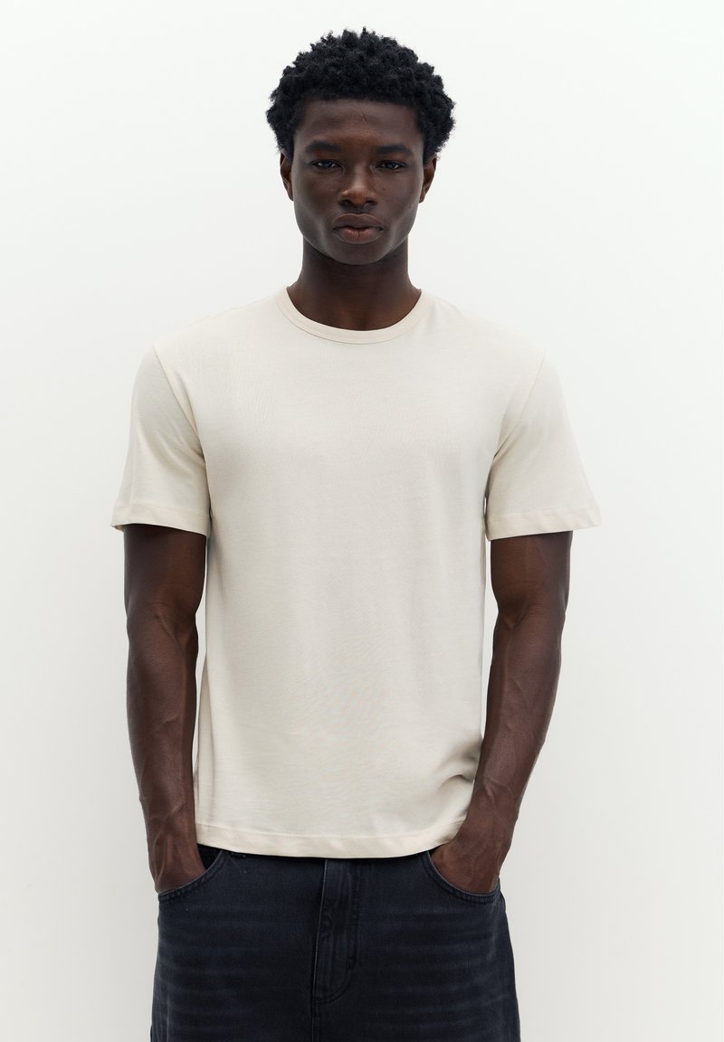 Jeune homme à la peau foncée et aux cheveux bouclés portant un t-shirt beige uni à manches courtes et un pantalon foncé, les mains dans les poches.