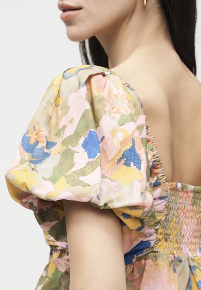 Γυναίκα που φοράει παστέλ λουλουδάτο φόρεμα με φουσκωτά μανίκια, κορδόνια στην πλάτη και χρυσό κρίκο στο αυτί, εμφανιζόμενη από το πλάι και από πίσω.