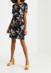 Robe à manches courtes florale en tissu bleu marine avec des motifs jaunes, roses et verts, dotée d'une taille à nouer et d'une coupe décontractée. Associée à des sandales à talons jaunes.