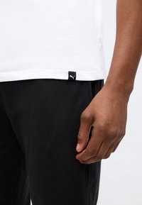 Egy kéz pihen fekete nadrág mellett, fehér inggel, amelynek alján egy kis Puma logós címke található, sima háttér előtt.