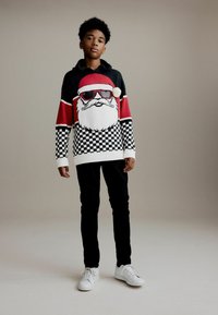 Zwart en rood gebreide trui met een Santa-gezicht met zonnebril, geruit patroon aan de onderkant, gecombineerd met zwarte broek en witte sneakers.