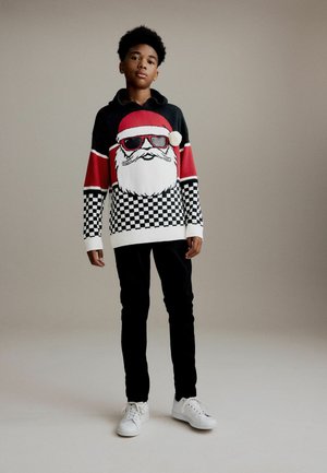 Pull en tricot noir et rouge avec un visage de Santa portant des lunettes de soleil, motif à carreaux en bas, associé à un pantalon noir et des baskets blanches.