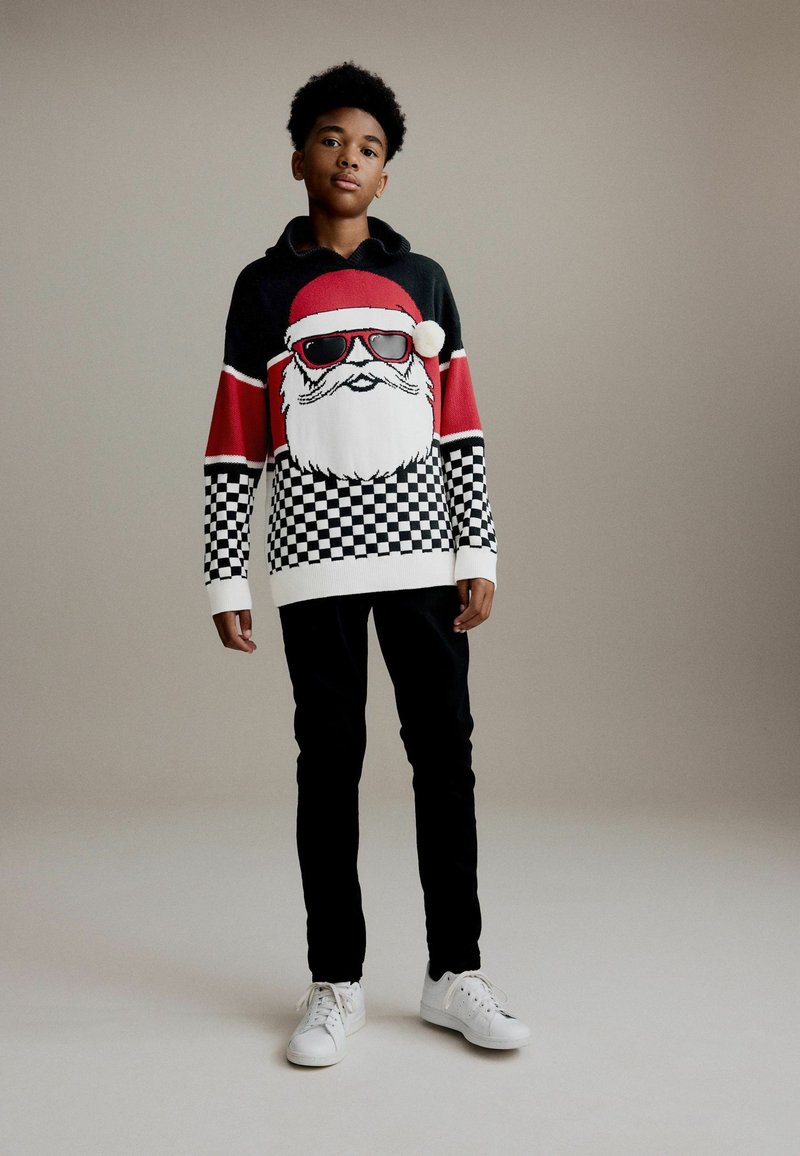 Zwart en rood gebreide trui met een Santa-gezicht met zonnebril, geruit patroon aan de onderkant, gecombineerd met zwarte broek en witte sneakers.