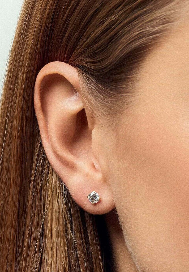 Söljdstudsörhänge med en klar rund ädelsten i en fyrprongsdesign, bärs i höger öra mot slät, rak hår.