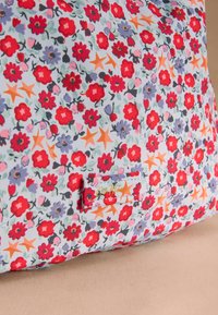 Cuscino in tessuto a motivo floreale con fiori rossi, viola e arancioni e foglie verdi su uno sfondo azzurro chiaro. Etichetta con il nome del marchio.