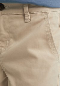 Pantaloni casual beige con tasche frontali visibili, passante per cintura e dettaglio del bottone, abbinati a una camicia azzurra infilata dentro.