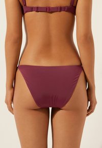 Bas de bikini en bourgogne profond, fabriqué à partir d'un matériau doux. Présente une coupe taille basse et une couverture minimale avec des coutures latérales.