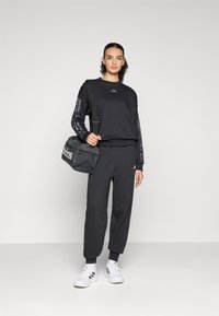 Donna in tuta Adidas nera e sneakers bianche, con una borsa da palestra Adidas nera a tracolla, su uno sfondo semplice.