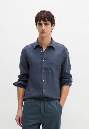 Młody mężczyzna ubrany w ciemnoniebieską koszulę zapinaną na guziki z podwiniętymi rękawami oraz pasujące spodnie ze sznurkiem, stojący na tle jednolitego białego tła.