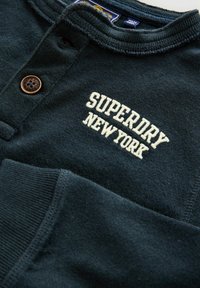Tume roheline dressipluus tekstureeritud pinnaga, millel on nööpidega kaelus ja kreemivärviline "SUPERDRY NEW YORK" tikand.