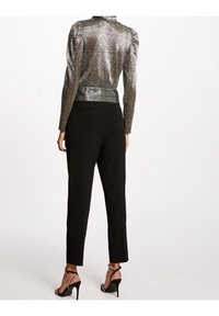 Frau, die ein metallisches Langarmoberteil, schwarze hochtaillierte Hosen und schwarze hochhackige Sandalen mit Knöchelriemen trägt, steht mit dem Rücken zugewandt.