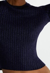 Gros plan sur une personne portant un haut tricoté ajusté, côtelé, pailleté bleu foncé avec des manches courtes.