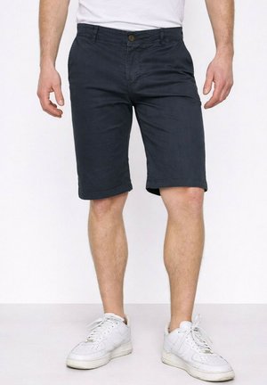Homme portant un short bleu marine foncé, des baskets blanches et un t-shirt blanc, debout devant un fond clair uni.