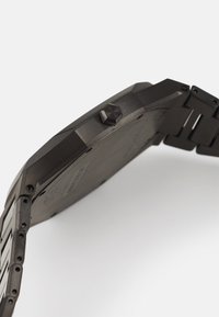 Montre noire en métal avec un boîtier octogonal et un bracelet texturé. Dispose d'une couronne proéminente et de détails matériels visibles. Fini lisse dans l'ensemble.