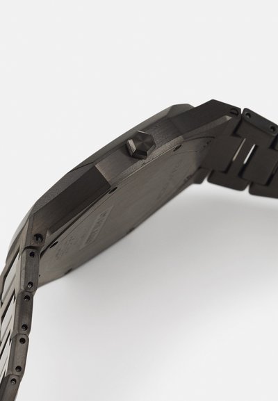 Μαύρο μεταλλικό ρολόι με οκταγωνική θήκη και υφασμάτινο λουρί. Διαθέτει έντονο κορώνα και ορατές λεπτομέρειες υλικών. Λείο φινίρισμα συνολικά.