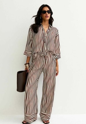 Mujer con camisa de manga larga abotonada de rayas verticales marrones y blancas y pantalones anchos a juego, sosteniendo un bolso marrón y con gafas de sol.