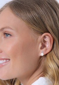 Boucles d'oreilles à clou en perle blanche avec un petit accent de gemme claire, montées sur un support en métal, présentées sur l'oreille d'un modèle à la peau claire.