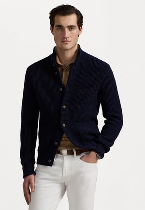 Cardigan in maglia blu navy con collo alto, caratterizzato da un motivo testurizzato, due tasche frontali e bottoni in legno, abbinato a una camicia beige e pantaloni bianchi.