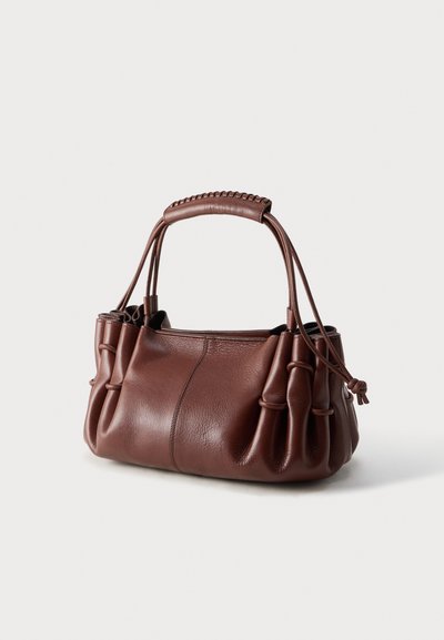 Καφέ δερμάτινη τσάντα με σούρες στα πλευρά, διπλές λαβές και ραμμένες λεπτομέρειες, με απλό ανοιχτό φόντο.