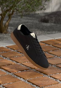 Zwarte leren sneaker met een gestructureerd oppervlak, voorzien van een beige hiel en veters, en een tan rubberen zool. Het logo "CK" is op de zijkant afgebeeld.