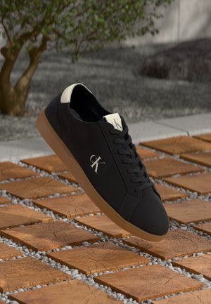 Zwarte leren sneaker met een gestructureerd oppervlak, voorzien van een beige hiel en veters, en een tan rubberen zool. Het logo "CK" is op de zijkant afgebeeld.