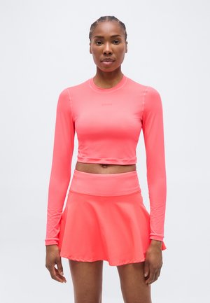 Femme debout, portant un haut court à manches longues corail vif et une jupe courte taille haute assortie, sur un fond blanc uni.