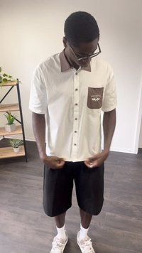 Homem a usar óculos, camisa bege abotoada com colarinho e bolso castanho, calções pretos, meias brancas e ténis, de pé no interior.