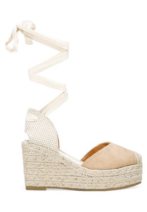 Sandalia de cuña con parte superior de ante beige, suela de yute trenzado y cordones de tela blanca en espiral subiendo por el tobillo. Diseño de punta abierta con un acento texturizado.