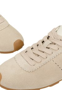 Béžové semišové tenisky s odpovídajícími tkaničkami, kulatou špičkou a světle hnědou gumovou podrážkou. Prošívání podél okrajů.