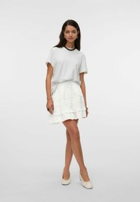 Top blanco de manga corta combinado con una falda blanca de volantes que presenta detalles texturizados. La modelo lleva zapatos beige y un collar negro como acento.