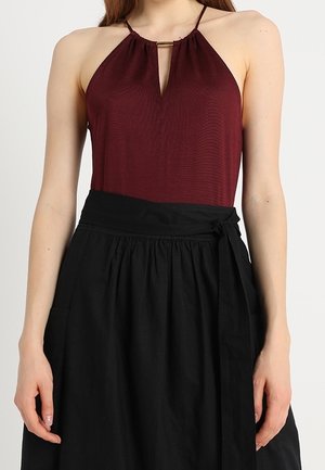 Kobieta ubrana w bordowy top na ramiączkach z wiązaniem na szyi oraz czarną spódnicę z wysokim stanem i wiązaniem po bokach, stojąca na jednolitym tle.