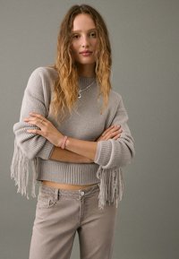 Cropped, ribbed, gray sweater with fringe detailing at sleeves, paired with light gray jeans. Model stands with arms crossed against a gray background.