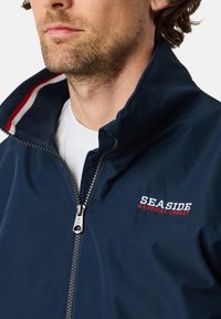 Marinejakke med opretstående krave, lynlås og rød-hvid stribet foer. Har broderet "SEASIDE NAUTICAL LEGACY" logo.