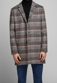 Cappotto a quadri marrone con accenti rosa, dotato di colletto a rever, chiusura con tre bottoni e taglio di lunghezza media. Indossato sopra un dolcevita nero.