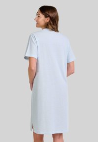 Mujer sonriendo, de perfil, vestida con un vestido corto de manga corta en azul claro que llega a la rodilla y con una pequeña abertura lateral, sobre un fondo gris.