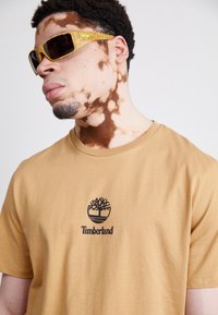 Beige bomull T-skjorte med en svart Timberland-logo på brystet, sammen med lyse solbriller med teksturert ramme.