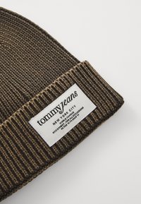 Beanie tricotat maro, cu textură în relief, având o etichetă albă cu text și logo negru. Design simplu, casual, pentru căldură.