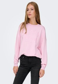 Sudadera rosa con bordado frontal "PARIS". Tejido suave, corte relajado, hombros caídos y puños acanalados. Combinada con jeans negros.