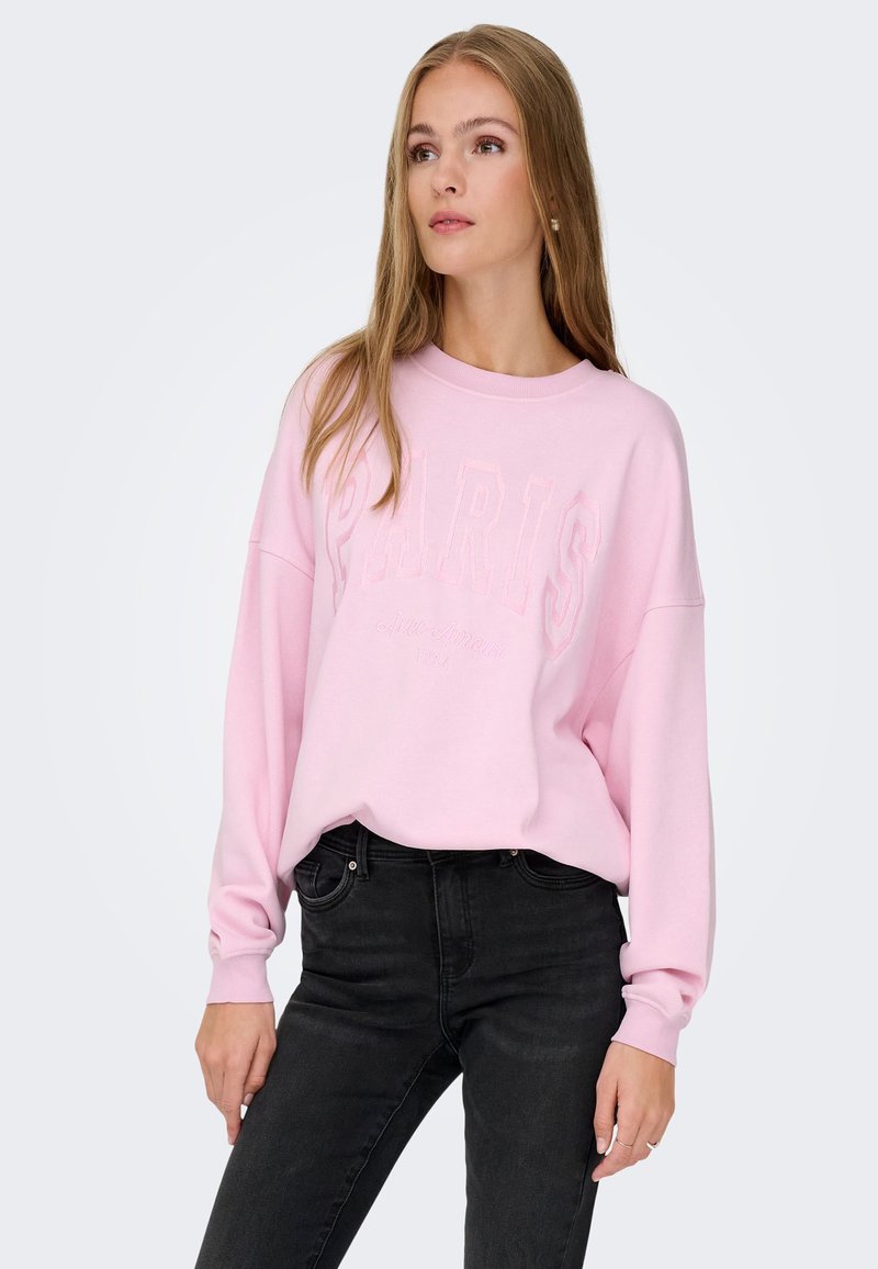 Sudadera rosa con bordado frontal "PARIS". Tejido suave, corte relajado, hombros caídos y puños acanalados. Combinada con jeans negros.