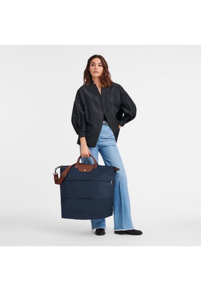 Navy puffer tote with brown leather accents. Large rectangular shape, zip closure. Model wears a black top and blue flared jeans.