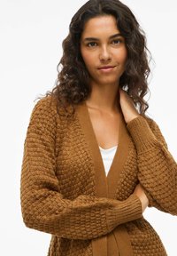 Cardigan en tricot marron avec un motif texturé, décolleté en V profond et poignets côtelés. Superposé sur un haut blanc, présentant une coupe décontractée.