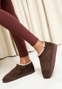 Mocassini in camoscio marrone con bordatura beige testurizzata, altezza bassa e suola piatta in gomma, abbinati a leggings color bordeaux.