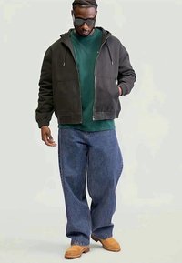Temna zip-up kapuca z teksturirano površino, čez zelen sveter. V kombinaciji z ohlapnimi modrimi kavbojkami in tan z velikimi suede čevlji. Elegantna sončna očala dopolnjujejo videz.