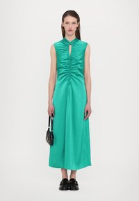 Satin teal zelena obleka z izrezom v obliki ključavnice in nabrano sprednjo stranjo, ki sega do sredine meče. Model drži majhno črno torbico in nosi usnjene čevlje.