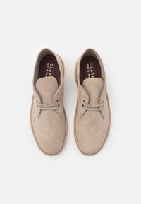 Zapatos de ante beige con punta redonda, que presentan una construcción ligera, cordones finos y una suela de color marrón claro contrastante.