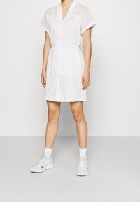 Kobieta ubrana w białą sukienkę na zamek błyskawiczny z krótkimi rękawami i bocznymi paskami, w białe skarpetki i białe sneakerzy, stojąca na tle jednolitym.