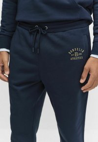 Man som bär marinblå träningsbyxor med dragsko och BonDeliD Athletics logotyp på låret, med händerna vila längs sidorna.